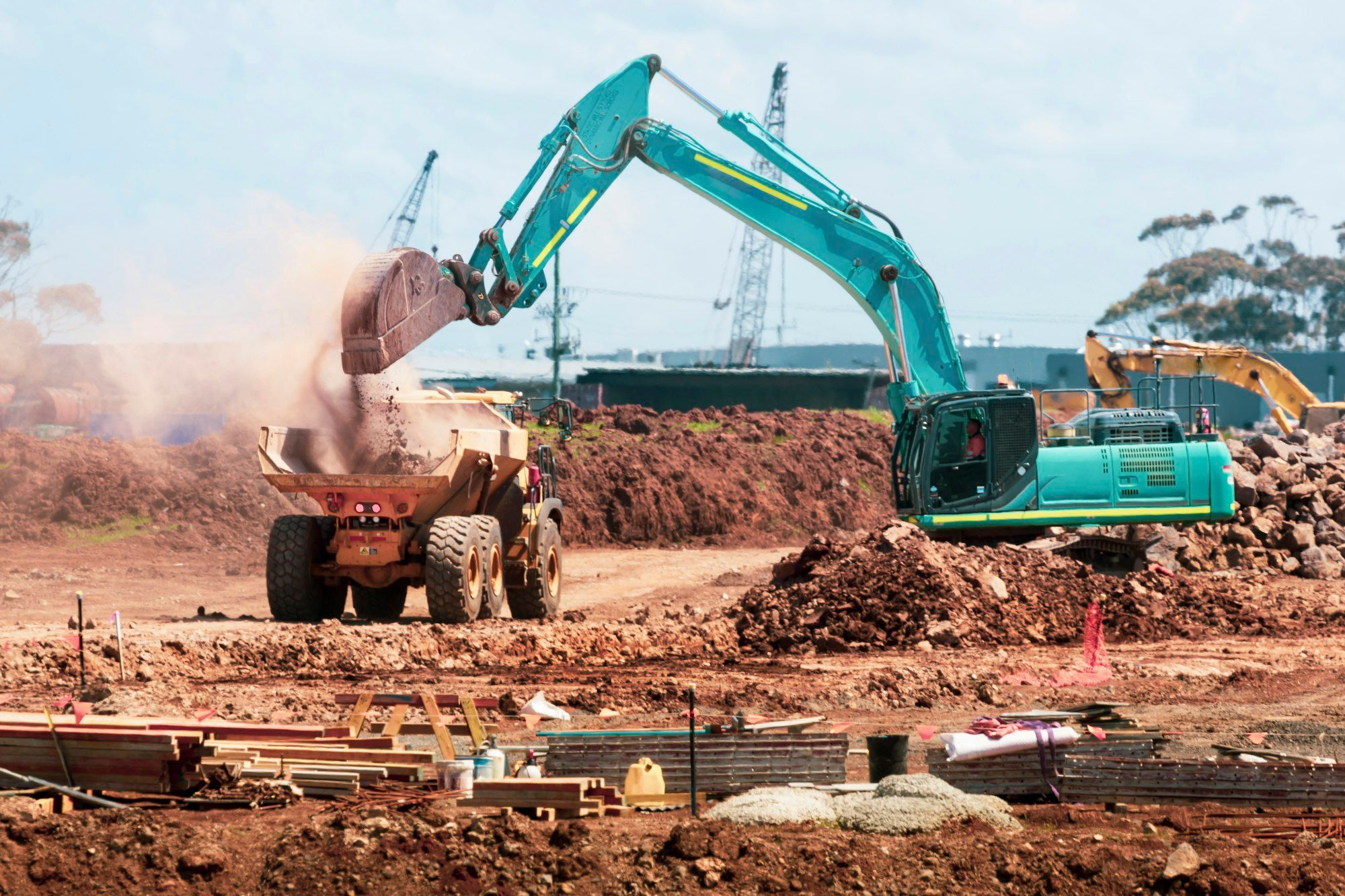 Teal excavator dumping soil into a yellow dump truck on a construction site