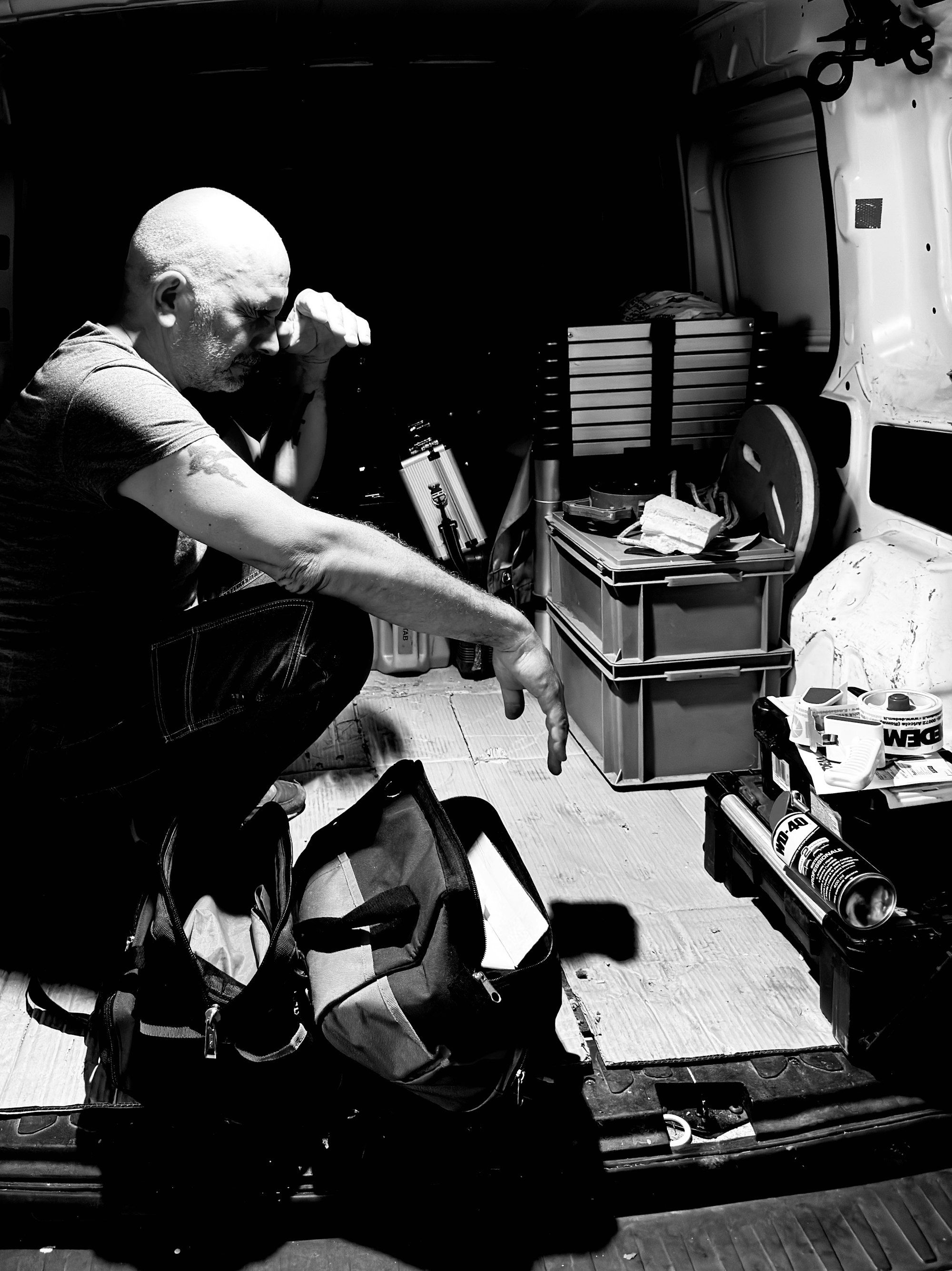 Tradesperson crouching in the back of a work van among tools, crates and a ladder